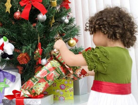 kisegyerek bontogatja az ajándékot a karácsonyfa előtt